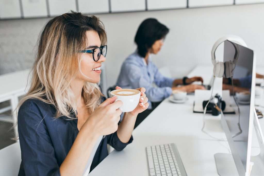 Female it-specialist looking at computer screen while drinking coffee with pleasure Indoor portrait of charming blonde secretary in glasses working beside asian male colleague | Fratello Coffee Roasters University Student